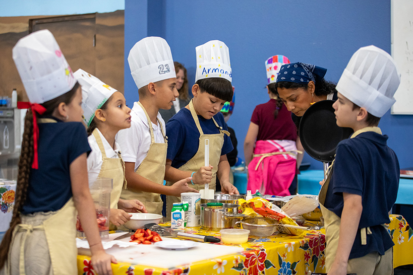 Students in aprons and chefs hats stand around a table preparing their dish