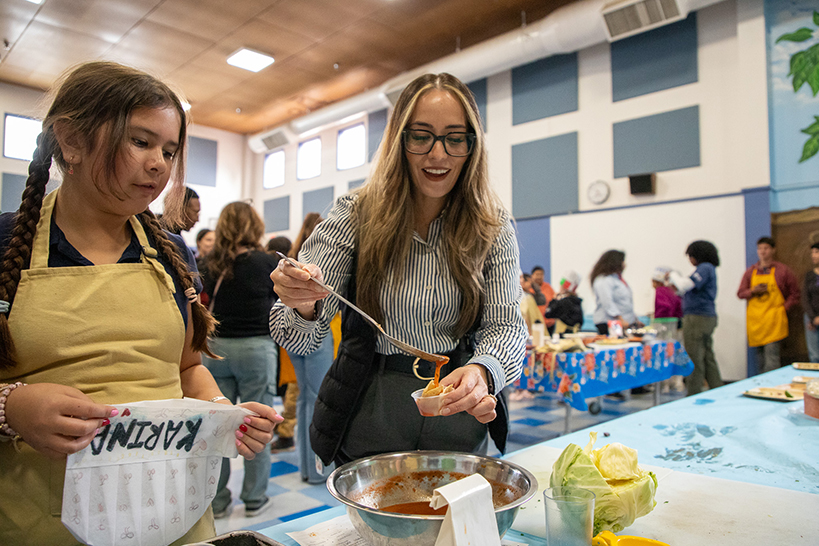 A woman spoons salsa on her cup of chips, as a young girl takes off her chef hat