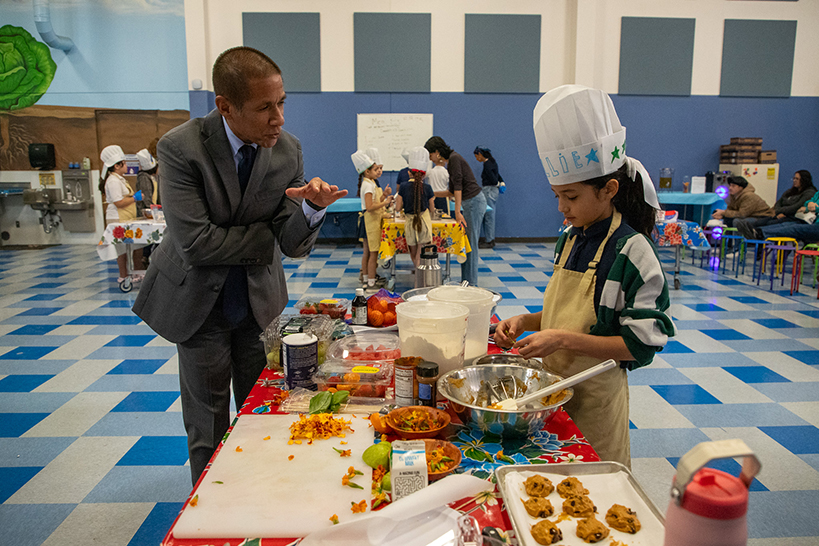 A man in a suit talks to a girl as she preps some sweet treats for the judges