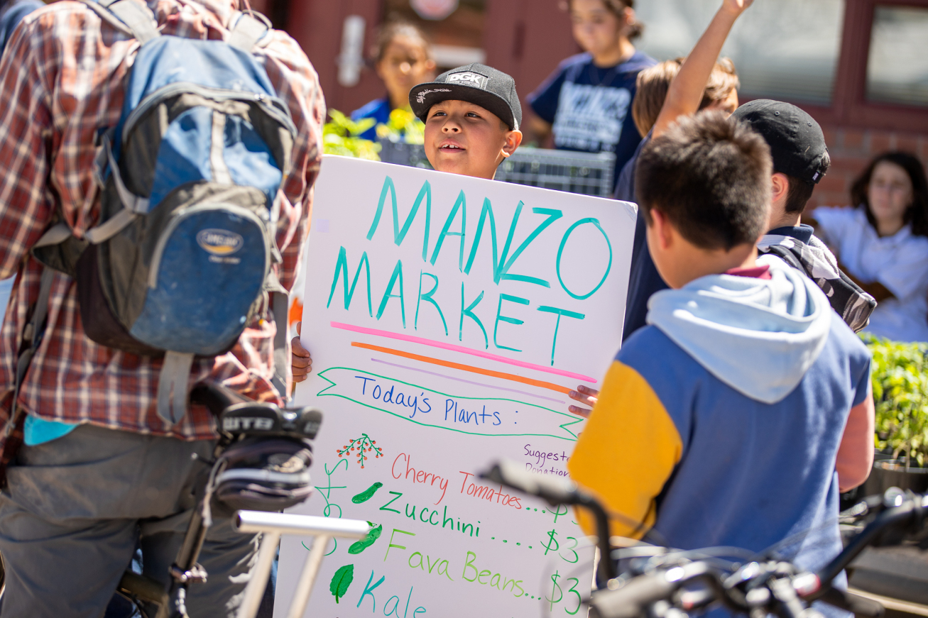 A boy holds up a sign reading Manzo Market with the plants of the day listed.